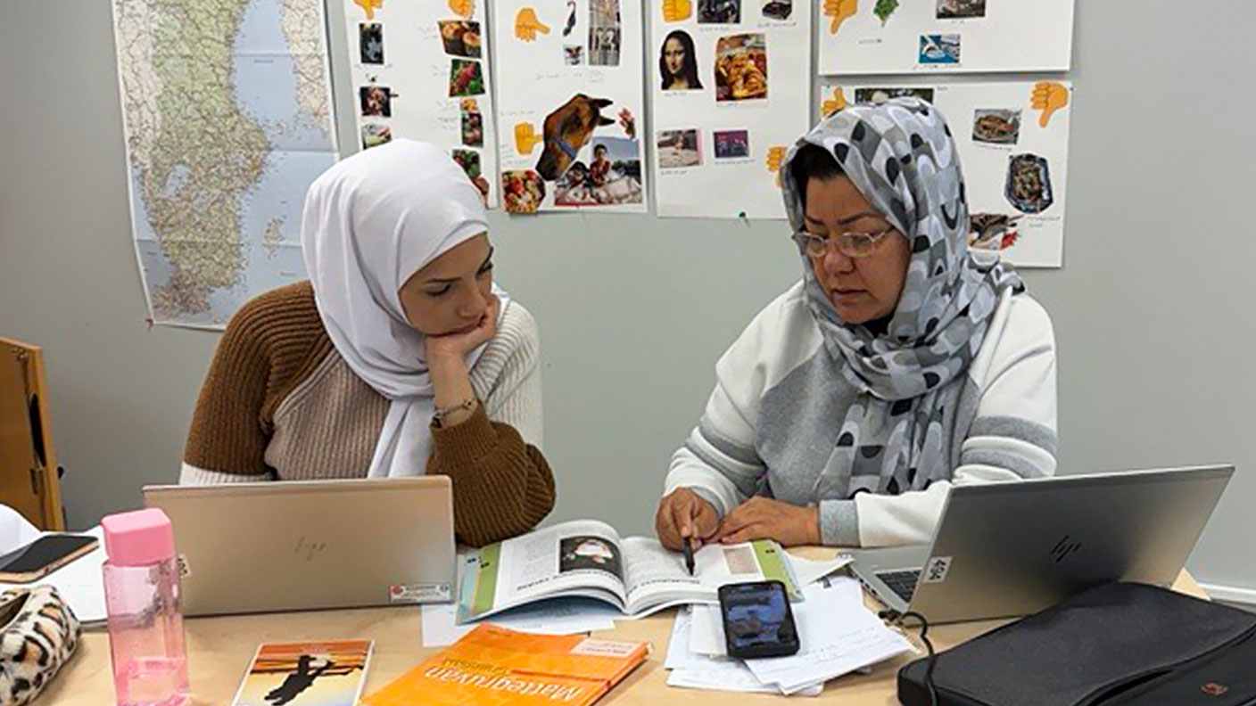 Två kvinnor med hijab sitter och studerar i skolbok vid bord med datorer, böcker och papper.