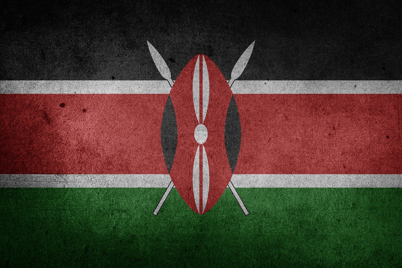 Kenyas flagga. Två vita korslagda spjut bakom oval sköld i rött, svart och vit. I botten vågräta färgfält, ovanifrån svart, rött och grönt, separerade med vita linjer. 