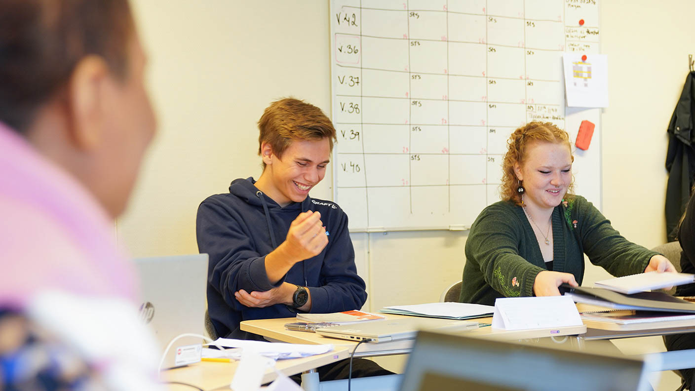 Två glada unga vuxna sitter vid skolbänk i klassrum.